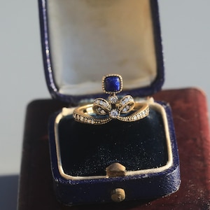 May include: A gold ring with a bow design, set with small diamonds and a square, dark blue gemstone. The ring is presented in a vintage blue jewellery box with a velvet interior.