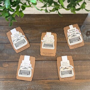 May include: Five magnetic bookmarks attached to brown cardstock backing, each featuring a vintage typewriter illustration adorned with floral accents and inspirational text including Psalm 139:14, Matthew 19:26, Psalm 46:10, 2 Peter 3:18, and God's not done writing your story.