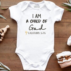 May include: White baby bodysuit with the text "I AM A CHILD OF God" and "Galatians 3:26". The bodysuit is surrounded by a wooden baby rattle, a pair of pink and white baby boots, and greenery on a wooden surface.