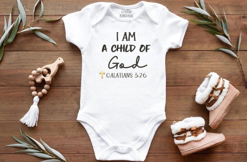 May include: White baby bodysuit with the text "I AM A CHILD OF God" and "Galatians 3:26". The bodysuit is surrounded by a wooden baby rattle, a pair of pink and white baby boots, and greenery on a wooden surface.