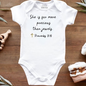 May include: White baby bodysuit with the text "She is far more precious than jewels. Proverbs 31:10." A gold cross is below the text. The bodysuit is on a wooden surface with baby shoes and a wooden toy.