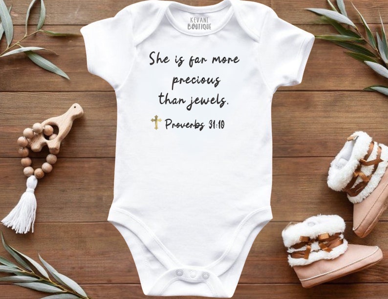 May include: White baby bodysuit with the text "She is far more precious than jewels. Proverbs 31:10." A gold cross is below the text. The bodysuit is on a wooden surface with baby shoes and a wooden toy.