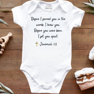 May include: White baby onesie with the text "Before I formed you in the womb. I knew you. Before you were born. I set you apart. Jeremiah 1:5." A gold cross is below the text. The onesie is next to a wooden rattle and baby boots.
