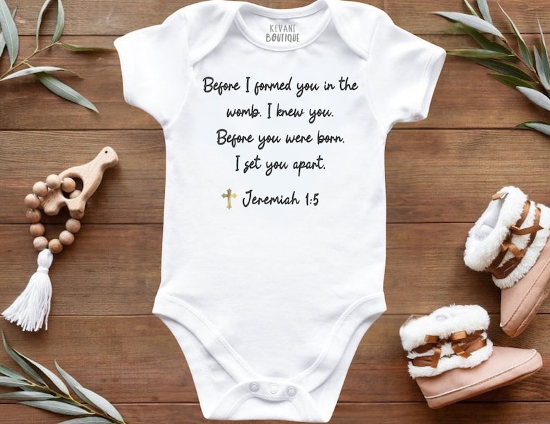 May include: White baby onesie with the text "Before I formed you in the womb. I knew you. Before you were born. I set you apart. Jeremiah 1:5." A gold cross is below the text. The onesie is next to a wooden rattle and baby boots.