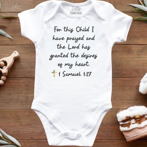 May include: White baby onesie with the text "For this Child I have prayed and the Lord has granted the desires of my heart. 1 Samuel 1:27." A gold cross is below the text. The onesie is next to a wooden rattle and baby boots.