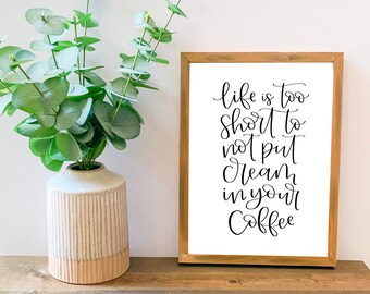 May include: A framed print with a white background and black calligraphy text that reads "Life is too short to not put cream in your coffee". The print is in a light brown wooden frame and is sitting on a wooden shelf with a vase of green eucalyptus leaves.