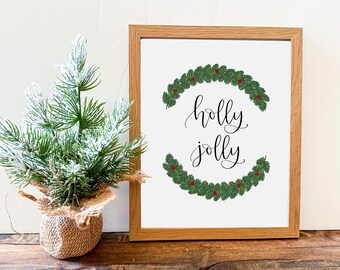 May include: A framed print with a white background and a green holly wreath with red berries. The text "holly jolly" is written in black cursive script inside the wreath.