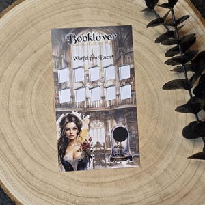 May include: A printed card with the word "Booklover" and the German phrase "W&uuml;rfel pro Buch!" The card features a woman holding a rose, blank book icons, and a library backdrop. A glass dome with a rose is also shown.