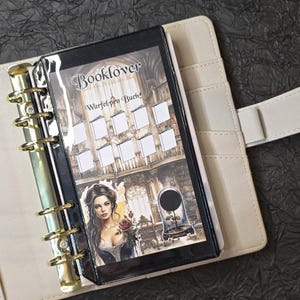 May include: A beige planner with gold rings, open to reveal a black insert with the word "Booklover" and the phrase "W&uuml;rfel pro Buch!" printed on it. The insert features a woman holding a rose and a library scene.