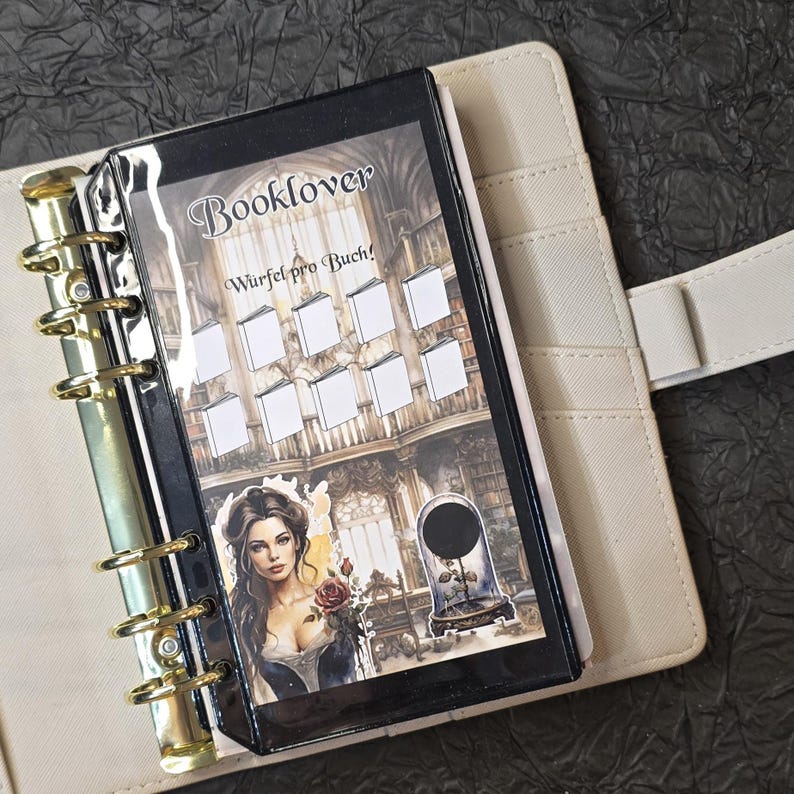 May include: A beige planner with gold rings, open to reveal a black insert with the word "Booklover" and the phrase "W&uuml;rfel pro Buch!" printed on it. The insert features a woman holding a rose and a library scene.