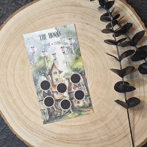 May include: A decorative card with the text "THE HOOD" and "PEEL = 11OS". The card features a watercolor illustration of a whimsical house, arrows, and small black circles. The card is set on a wooden surface.