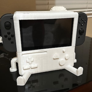 May include: White 3D printed stand for a Nintendo Switch console. The stand has a retro video game console design with buttons and a joystick. The Switch console is in the stand with a black Joy-Con controller attached to the left side.
