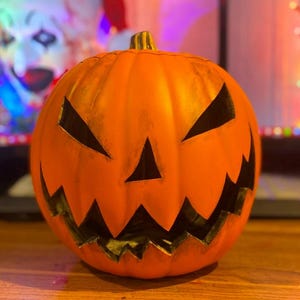 Puede incluir: Una calabaza naranja tallada con una cara de miedo. La calabaza tiene una amplia sonrisa con dientes y ojos negros.
