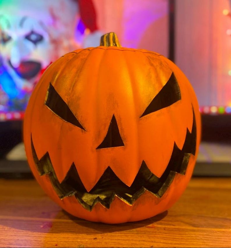 Puede incluir: Una calabaza naranja tallada con una cara de miedo. La calabaza tiene una amplia sonrisa con dientes y ojos negros.
