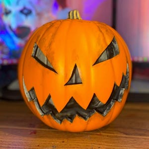 Puede incluir: Una calabaza naranja tallada con una cara de miedo. La calabaza tiene una boca, ojos y nariz negros.