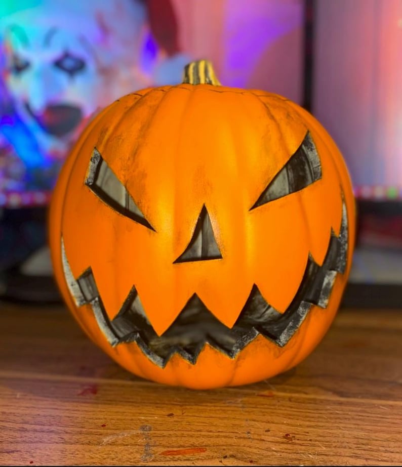 Puede incluir: Una calabaza naranja tallada con una cara de miedo. La calabaza tiene una boca, ojos y nariz negros.