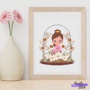 May include: A cross-stitch pattern featuring a cartoon princess wearing a pink dress and a gold crown, standing inside a glass dome with white flowers and brown branches.