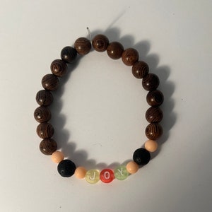 May include: A beaded bracelet featuring dark brown wooden beads and colorful accent beads. The word "JOY" is spelled out with letter beads in yellow, red, and green. The bracelet is strung on a clear elastic cord.