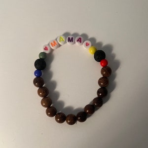 May include: A bracelet with the word "MAMA" spelled out in colourful letter beads. The bracelet features wooden beads, lava beads, and other colourful beads. The bracelet is on a white surface.