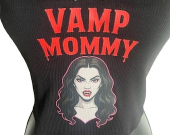 May include: A black ribbed crop top featuring a small black bow at the center neckline. The text VAMP MOMMY is printed in red above a graphic illustration of a woman with dark hair, pale skin, and visible fangs, outlined in a thin pink line.