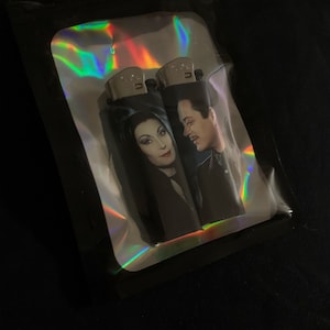May include: Two black lighters with a holographic finish. The lighters feature a printed image of Morticia and Gomez Addams from the Addams Family.