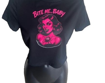 May include: A black short-sleeved crop top featuring a graphic print of a person with dark hair and fangs, accompanied by a spider illustration on the chest and the text "BITE ME, BABY" in bright pink lettering above the graphic design.