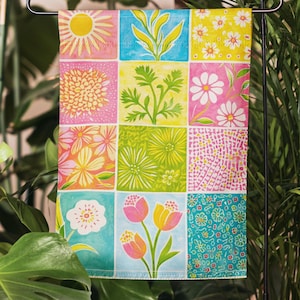 May include: A colorful garden flag featuring a grid of floral designs. The flag showcases various flowers, including daisies and tulips, in a vibrant palette of pink, yellow, blue, and green. The flag is displayed on a black metal stand amidst lush greenery.