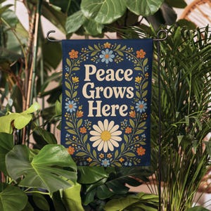 May include: A rectangular garden flag with a dark blue background featuring the text Peace Grows Here in cream letters. The design is framed by a symmetrical botanical border of green leaves and small orange, yellow, and light blue flowers.