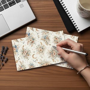 May include: Stationery set featuring a floral design with sunflowers, feathers, and crescent moons in shades of beige, blue, and brown. The stationery is on a wooden surface with a laptop and notebook in the background.