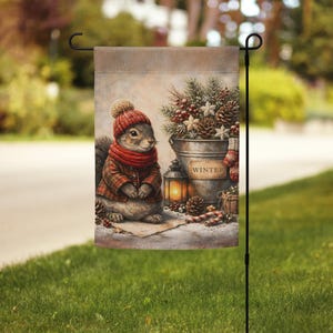 May include: A garden flag with a watercolour illustration of a squirrel in winter clothing. The squirrel is wearing a red hat, scarf, and coat. A metal bucket with pine cones and berries, a lantern, and the word "WINTER" are also shown.