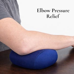 Puede incluir: Un coj&iacute;n ovalado azul marino que sostiene un codo. El coj&iacute;n est&aacute; sobre una superficie de madera marr&oacute;n oscuro. El texto "Elbow Pressure Relief" es visible en el fondo. La imagen muestra un brazo.