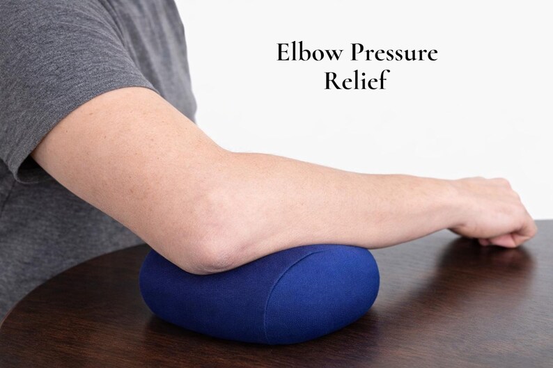 Puede incluir: Un coj&iacute;n ovalado azul marino que sostiene un codo. El coj&iacute;n est&aacute; sobre una superficie de madera marr&oacute;n oscuro. El texto "Elbow Pressure Relief" es visible en el fondo. La imagen muestra un brazo.