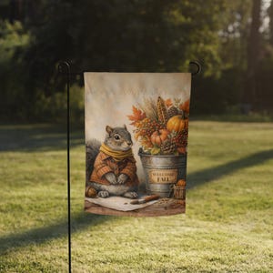 May include: A garden flag featuring a watercolor illustration of a squirrel wearing a scarf and jacket, with a bucket of fall harvest items. The bucket has a sign that says "WELCOME FALL". The flag is displayed on a black metal stand in a grassy yard.