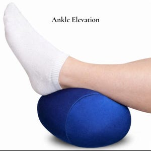 Puede incluir: Una almohada azul ovalada para la elevaci&oacute;n del tobillo. El texto "Ankle Elevation" est&aacute; encima de la almohada. Un pie con un calcet&iacute;n blanco descansa sobre la almohada.