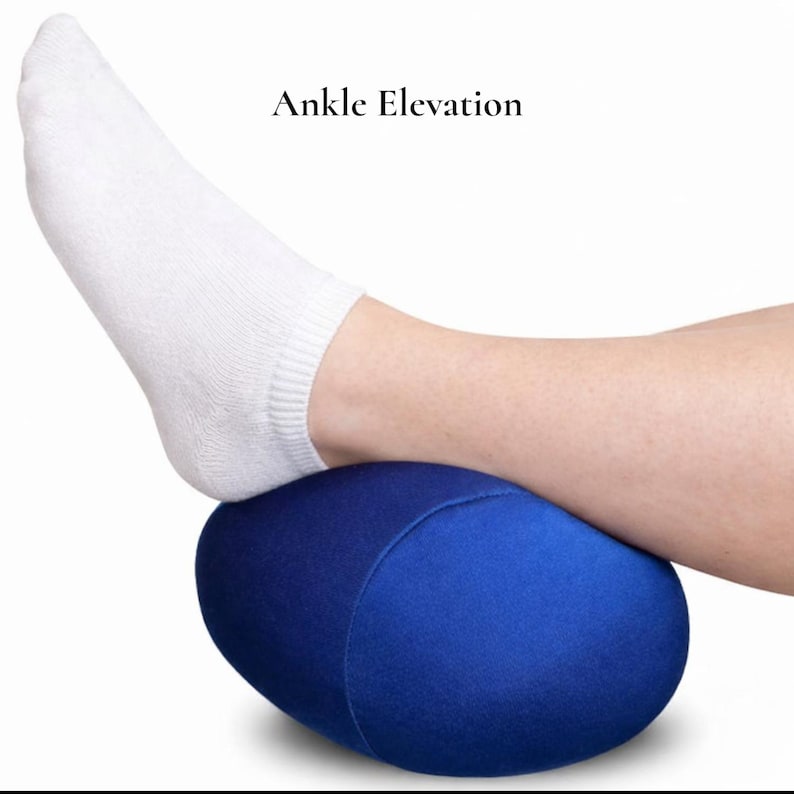 Puede incluir: Una almohada azul ovalada para la elevaci&oacute;n del tobillo. El texto "Ankle Elevation" est&aacute; encima de la almohada. Un pie con un calcet&iacute;n blanco descansa sobre la almohada.