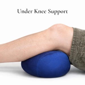 Puede incluir: Una almohada azul, de forma ovalada, para soporte debajo de la rodilla. La almohada est&aacute; debajo de la rodilla de una persona, brindando soporte. El texto "Under Knee Support" est&aacute; encima de la rodilla.