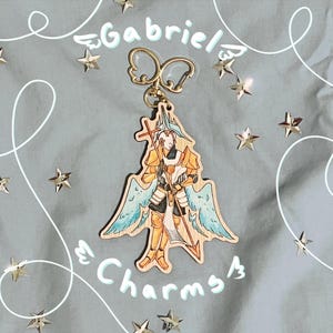 Puede incluir: Un llavero de madera con una ilustración impresa de una figura alada con armadura dorada que sostiene un bastón con una cruz. El dije tiene un cierre de metal dorado con forma de alas. El texto Gabriel Charms está escrito en azul claro.