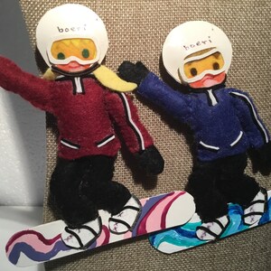 May include: Two felt figures of skiers wearing white helmets with the word "boeri" written on them. The skiers are wearing red and blue ski jackets and black pants. They are standing on painted wooden snowboards with pink, blue, and white designs.