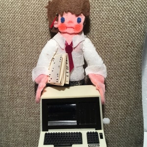 May include: A felt doll dressed in a white shirt and red tie, holding a stack of papers and standing in front of a toy computer with a black screen and a white keyboard.
