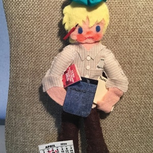 Könnte beinhalten: Handgefertigte Filzpuppe, die eine Person mit gestresstem Gesichtsausdruck darstellt, die ein Hemd, eine braune Hose und ein blaues Kopftuch trägt. Die Puppe hält ein Buch mit der Aufschrift "IRS" und ein Papier mit der Aufschrift "Taxes". Ein Kalender zeigt den Monat April 1900.
