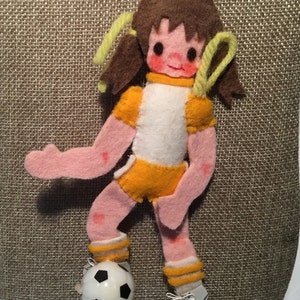 May include: A felt doll wearing a white and yellow soccer uniform, kicking a black and white soccer ball. The doll has brown hair with yellow pigtails and rosy cheeks.