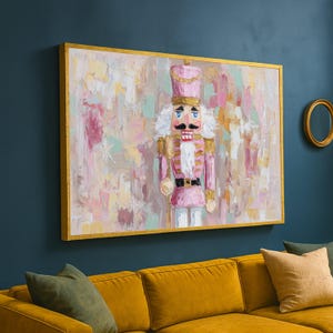 Può includere: Un'opera d'arte su tela incorniciata con un soldato schiaccianoci rosa con una cornice dorata. Il dipinto ha uno stile impressionista strutturato con una tavolozza di colori pastello di rosa, bianco e oro. L'opera d'arte è esposta sopra un divano giallo.