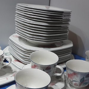 May include: A set of white porcelain teacups and saucers with a floral design. The cups and saucers have a delicate floral pattern in shades of pink, green, and brown. The set includes a stack of plates with a similar floral design.