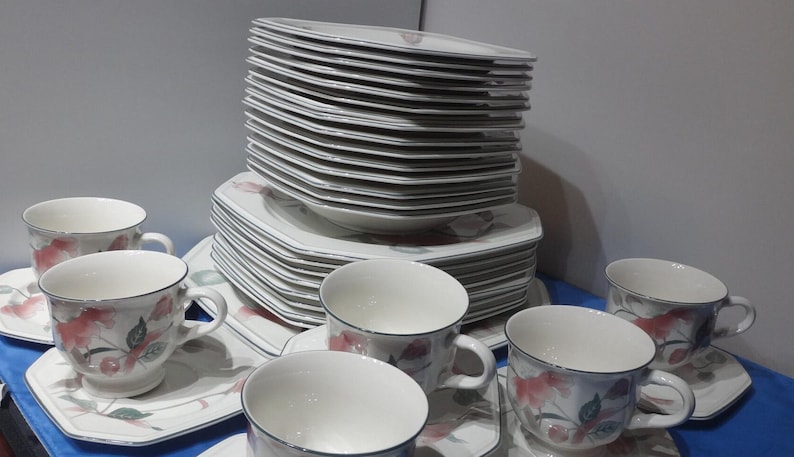 May include: A set of white porcelain teacups and saucers with a floral design. The cups and saucers have a delicate floral pattern in shades of pink, green, and brown. The set includes a stack of plates with a similar floral design.