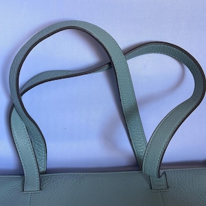 May include: A light blue tote bag with two curved handles. The handles are a lighter shade of blue with a darker trim. The bag has a textured surface and is made of a soft material.