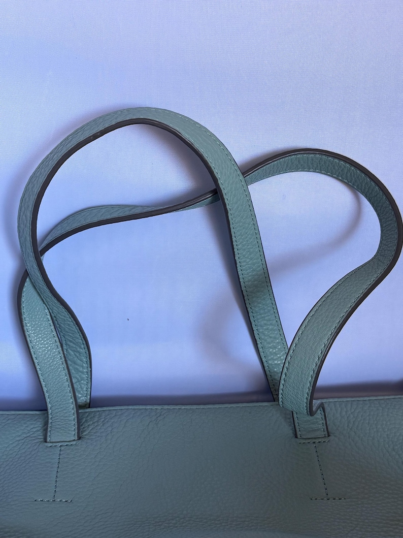 May include: A light blue tote bag with two curved handles. The handles are a lighter shade of blue with a darker trim. The bag has a textured surface and is made of a soft material.