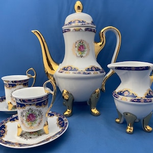 Può includere: Un servizio da tè in porcellana bianca con dettagli dorati, composto da una teiera, una lattiera, una zuccheriera e due tazze da tè con piattini. Il set presenta un motivo floreale e bordi blu.