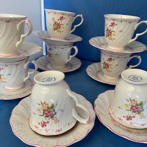May include: Set of six white teacups and saucers with pink floral patterns and gold accents. The cups and saucers have a scalloped edge.