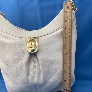 May include: Cream-colored leather shoulder bag with a gold-tone clasp and hardware. The bag has a curved shape and a single shoulder strap. The ruler next to the bag shows a length of approximately 12 inches.