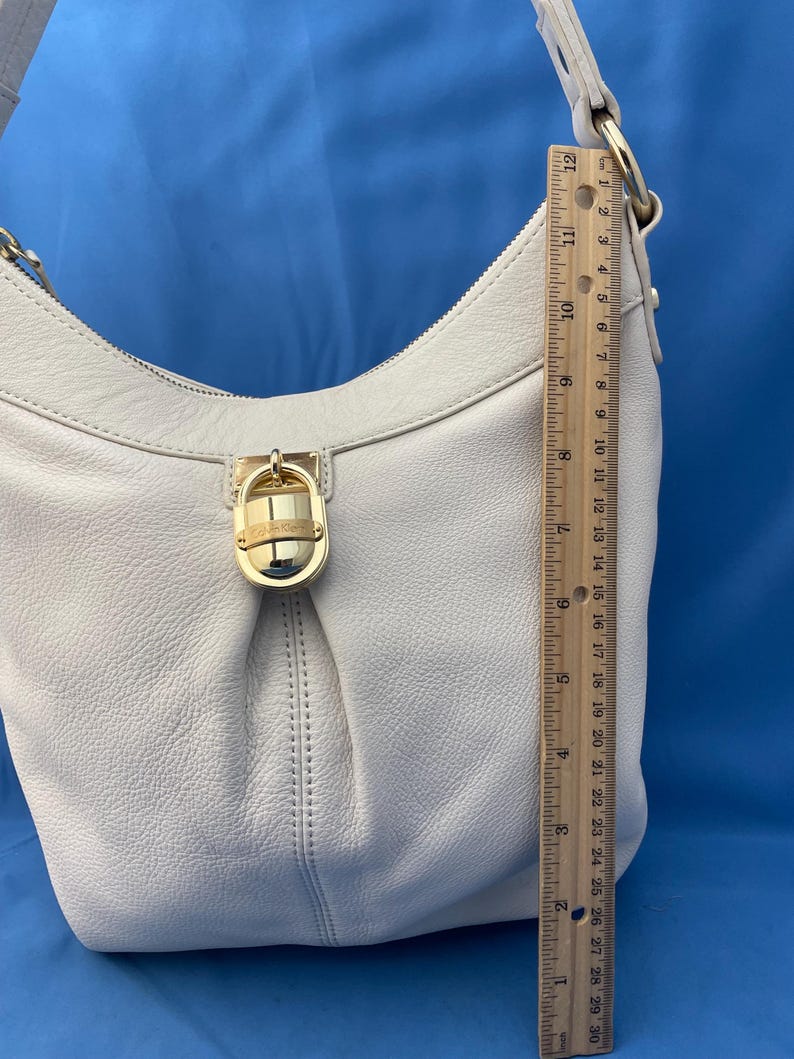 May include: Cream-colored leather shoulder bag with a gold-tone clasp and hardware. The bag has a curved shape and a single shoulder strap. The ruler next to the bag shows a length of approximately 12 inches.